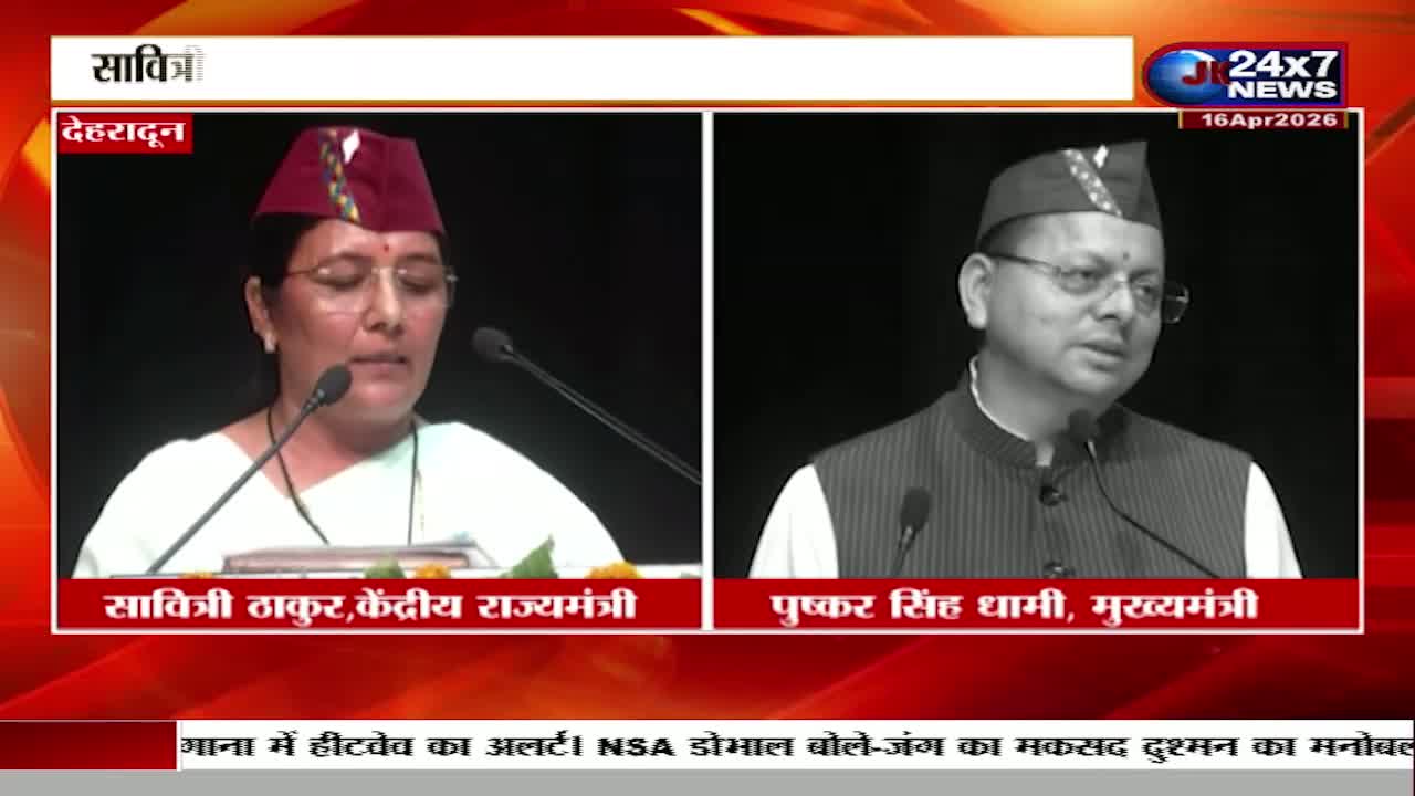 A woman in a maroon cap speaks into a microphone, her expression serious. Beside her, a man in a similar cap and vest addresses the audience.
