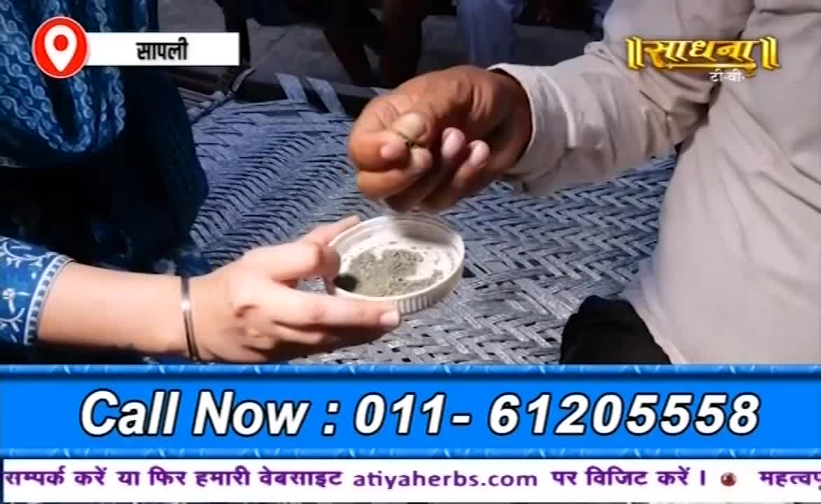 A man carefully pinches something from his fingers, dropping it into a small white container held by a woman. The Sadhna logo and a phone number flash on the screen below.

