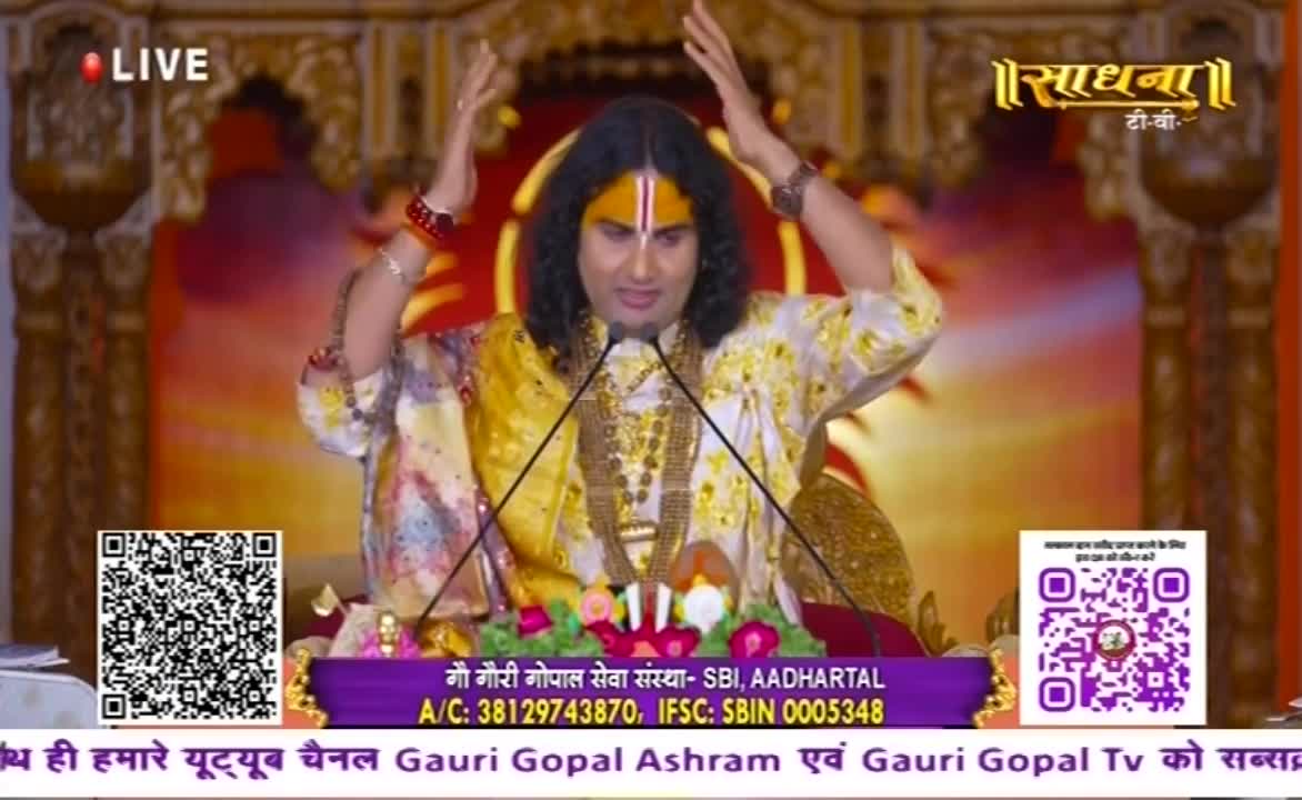 A man in a gold-patterned robe stands before two microphones, his arms raised. He's wearing a yellow mark on his forehead, and the Sadhna logo is visible in the background.
