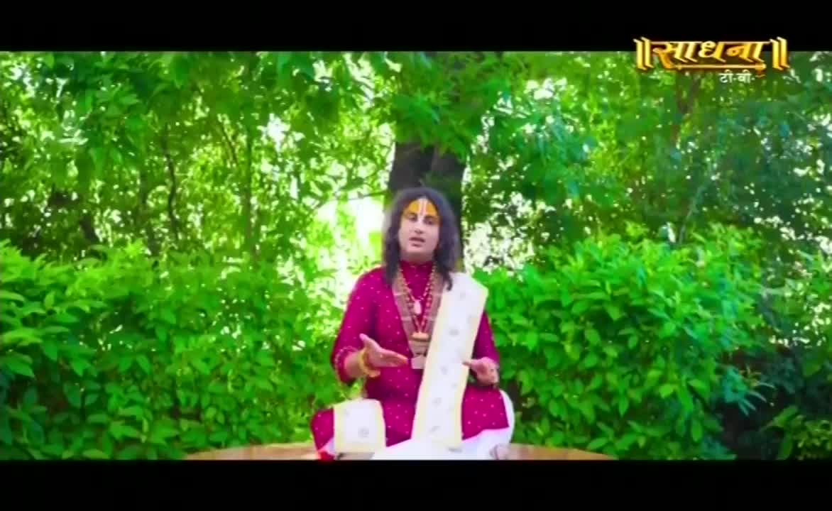 A man in a maroon kurta sits cross-legged, gesturing with his hands. He speaks, framed by lush green foliage, the Sadhna TV logo visible above.
