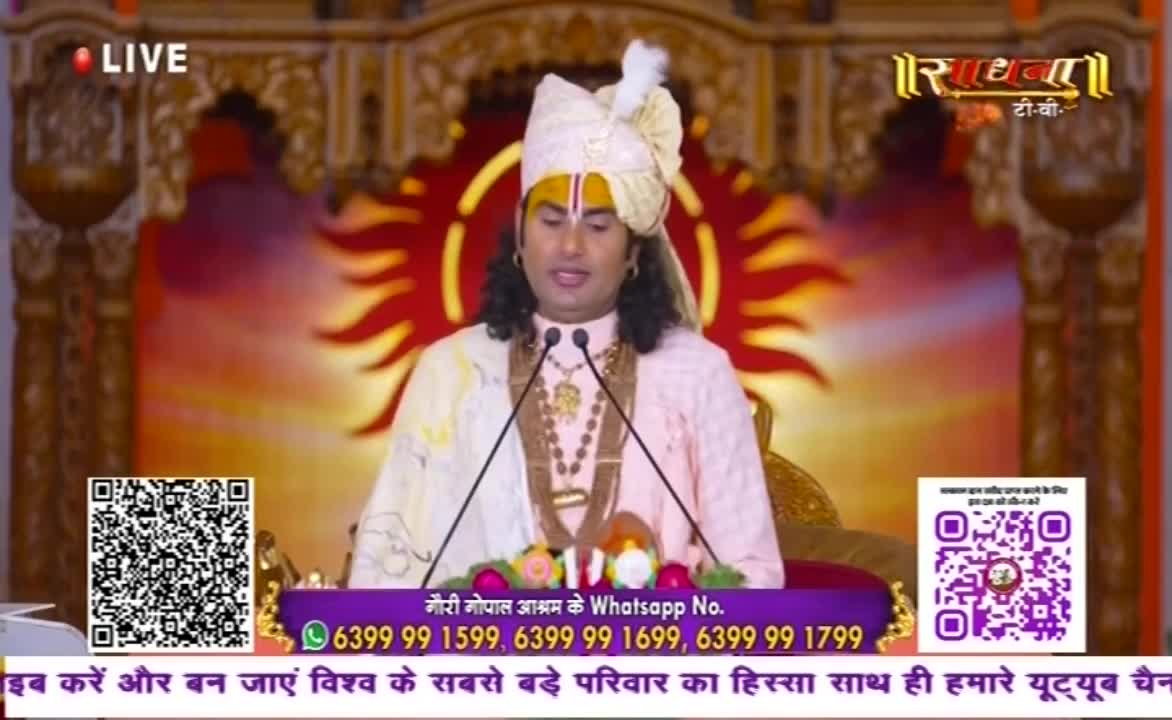 A man in a turban and ornate jewelry speaks into two microphones. The backdrop is a stylized, sun-like design, and the broadcast is live on Sadhna TV.

