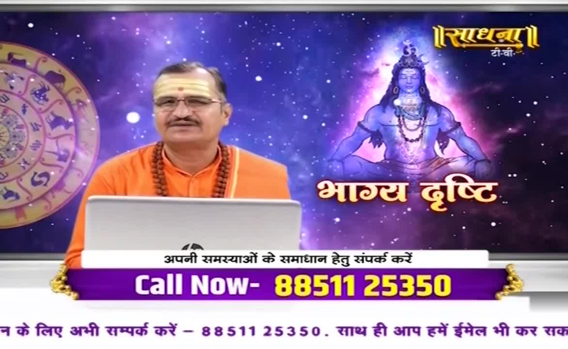 A man in saffron robes sits at a desk, a laptop open in front of him. Behind him, the Sadhna TV logo and an image of a deity are visible, alongside a phone number for viewers to call.
