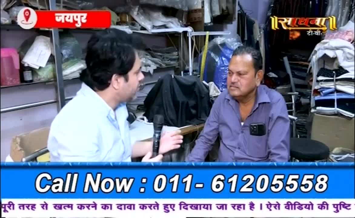 A man in a light blue shirt gestures with a microphone, speaking to a man in a purple shirt. The backdrop is a cluttered shop, with shelves and various fabrics visible.
