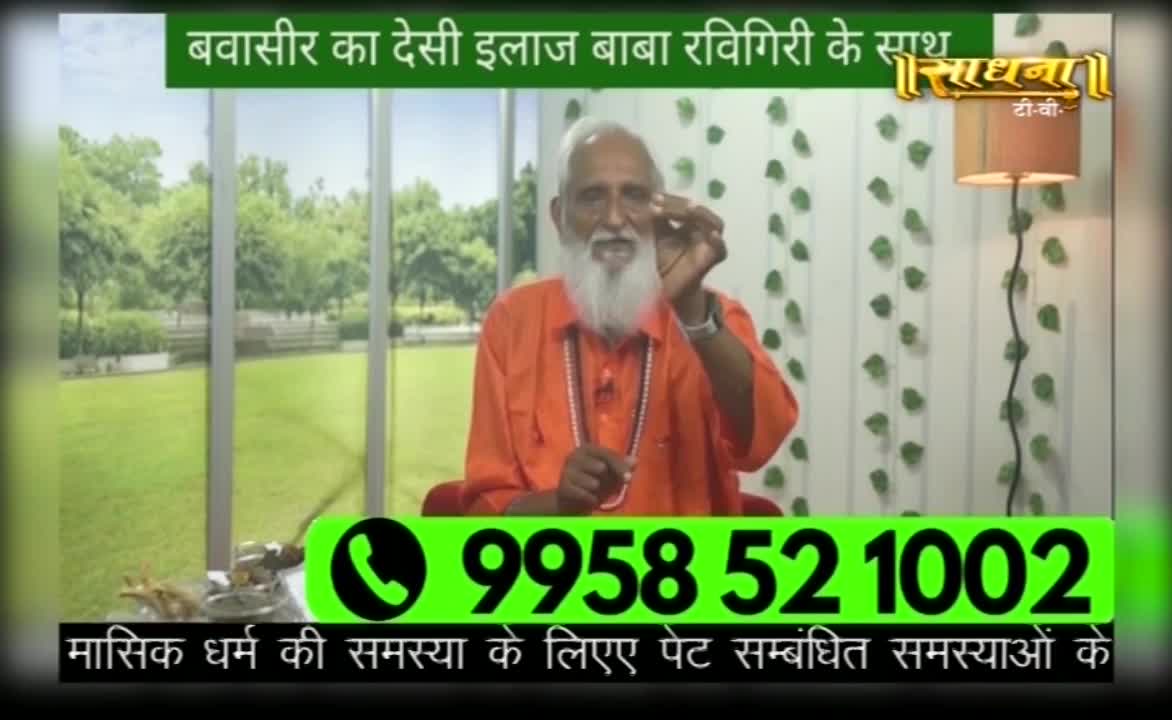 A man in an orange robe gestures with his hand while speaking on a show for Sadhna TV. Below him, a phone number is displayed in bright green, and the screen also has text in Hindi.
