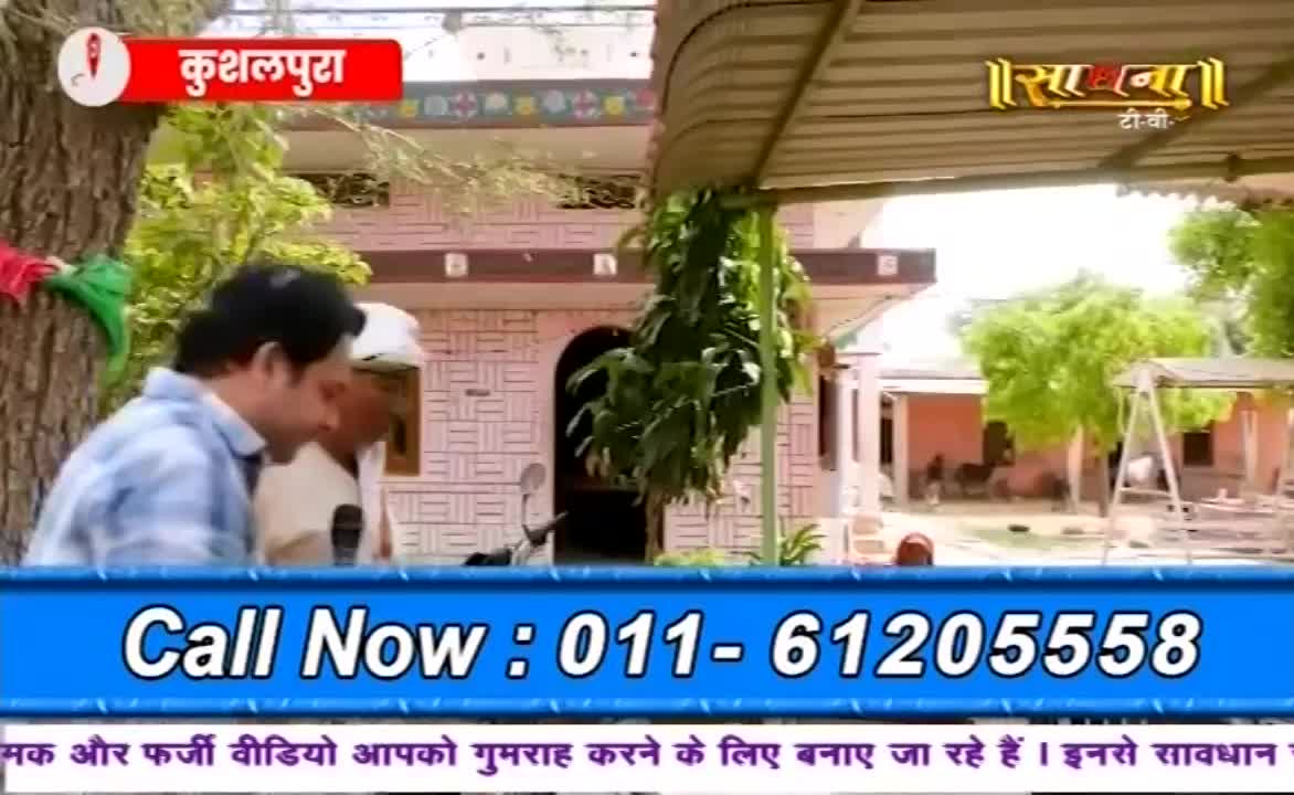 Two men stand outside a pink building, one holding a microphone, likely for a Sadhna TV broadcast. A phone number flashes on a blue banner, with text in Hindi warning about misleading videos.
