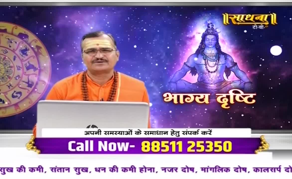 A man in saffron robes sits behind a laptop, speaking on a television program called Sadhna. Behind him, a celestial image of a deity is displayed, and a phone number flashes on the screen.
