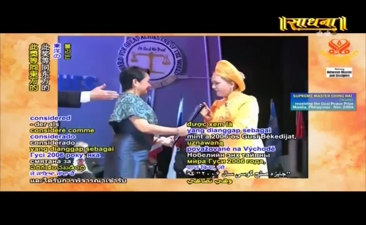 A woman in a yellow outfit presents a wrapped object to another woman, who's wearing a patterned shawl. The Sadhna logo and other text are visible on the screen behind them.
