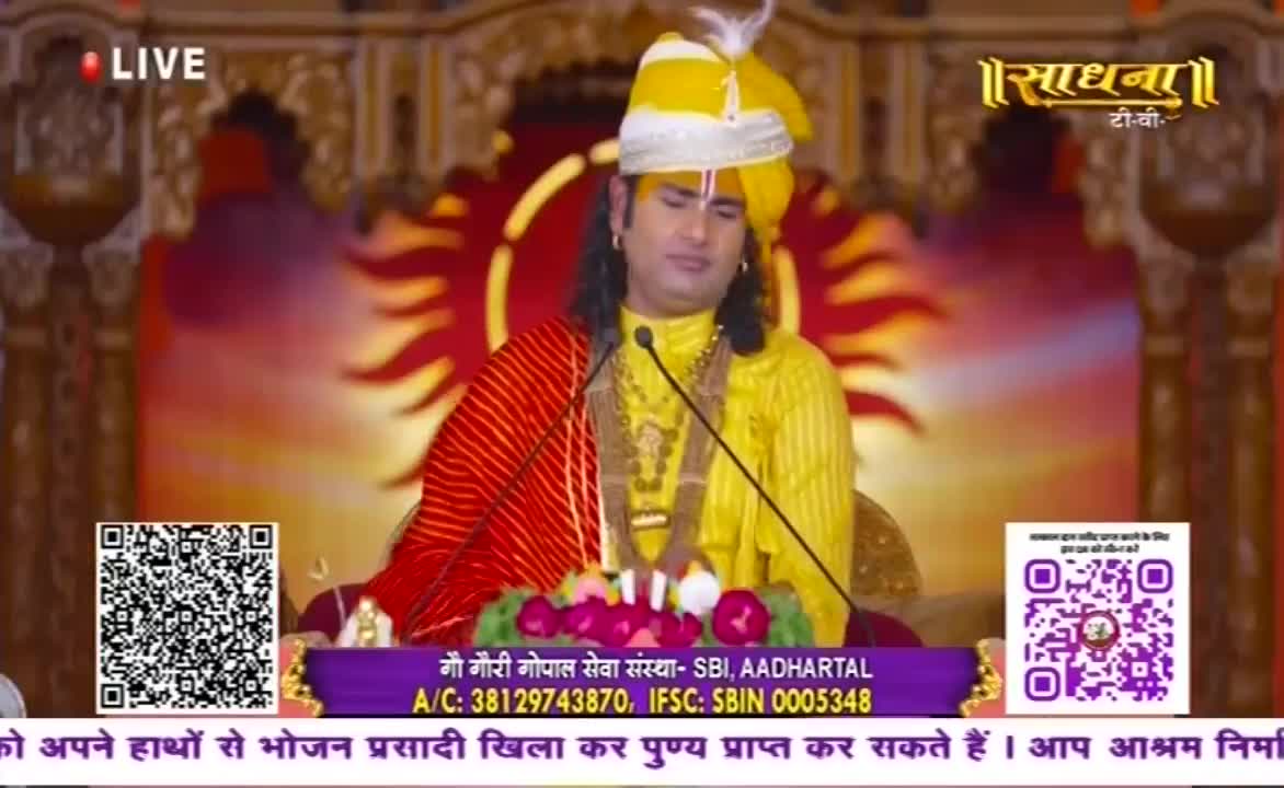A man in a yellow outfit and turban sits at a lectern, speaking into a microphone. The Sadhna TV logo is visible, and the broadcast is marked as "LIVE."
