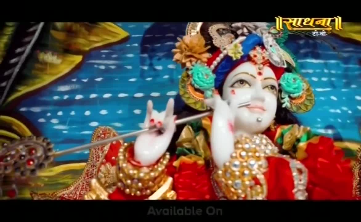 A statue of Krishna, adorned in jewels and a floral headdress, holds a flute. The background is a painted scene of water and sky, and the Sadhna logo sits in the corner.
