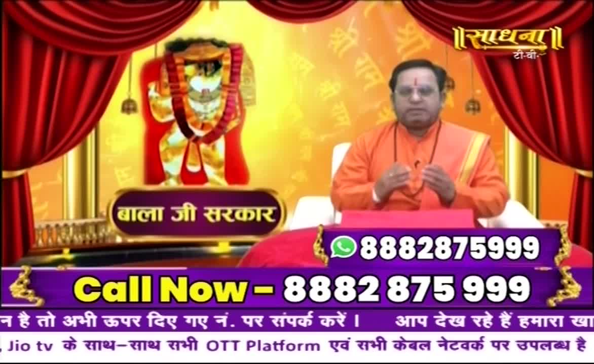 A man in an orange robe sits before a backdrop featuring an image of a deity, with a phone number displayed prominently below. The Sadhna TV logo is visible in the top right corner, and the words "Call Now" are displayed in a purple banner.
