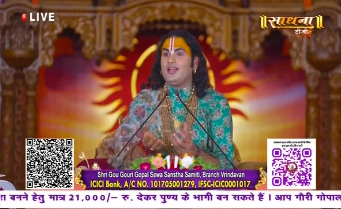 A man in ornate clothing, a yellow mark on his forehead, speaks animatedly into two microphones. The Sadhna TV logo and a "LIVE" indicator are visible, along with text about a charitable donation.
