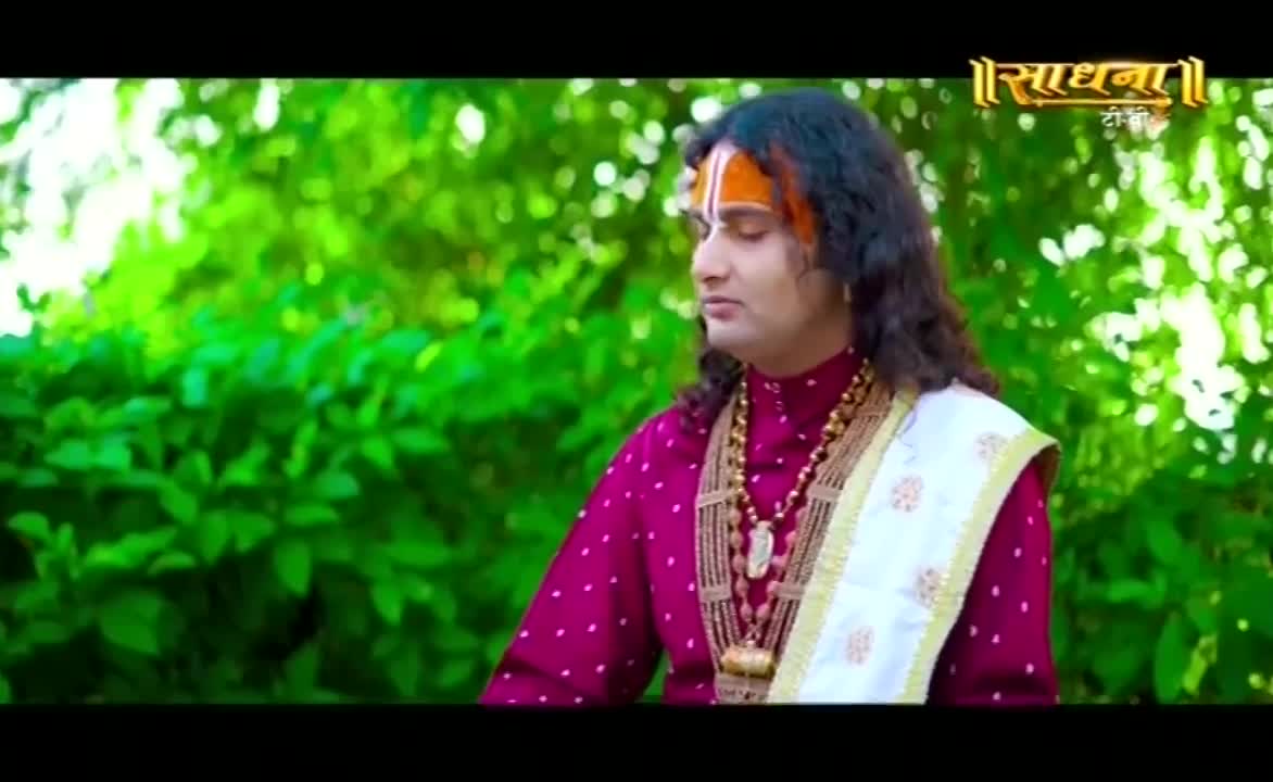 He's dressed in a maroon kurta, a white shawl draped over his shoulder, and a prominent tilak on his forehead. The man, seemingly in deep thought, is set against a backdrop of lush green foliage.
