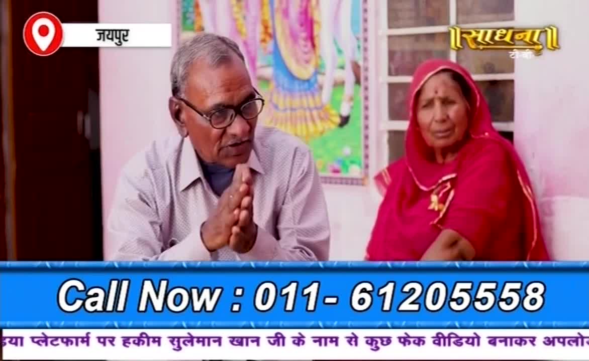 An older man with glasses and clasped hands speaks intently, while a woman in a red head covering looks on. The Sadhna logo and a phone number are displayed in the lower part of the screen.
