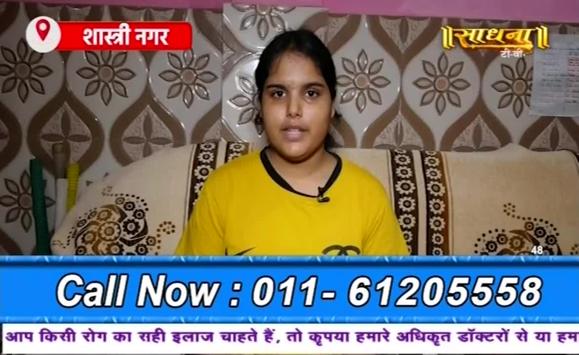 A young woman in a yellow shirt speaks directly to the camera, framed by a sofa and a wall with a repeating pattern. Below her, a blue banner displays a phone number, with text in Hindi.
