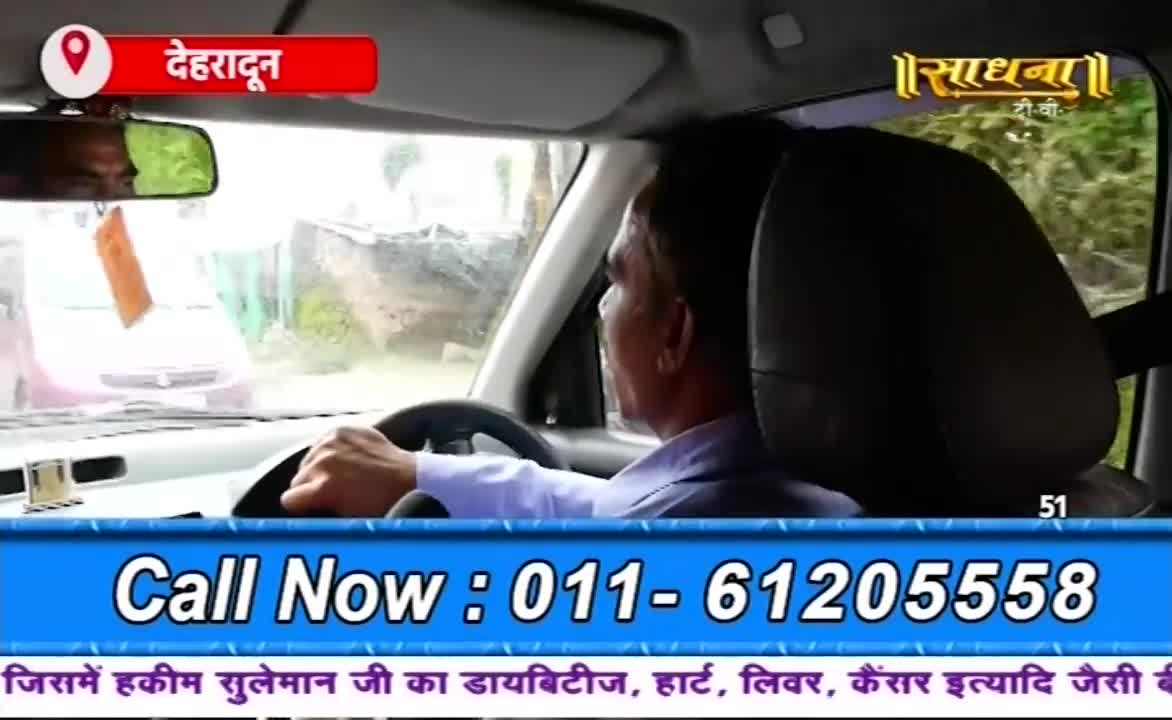 A man in a light blue shirt grips the steering wheel, his gaze fixed on the road ahead. The Sadhna TV logo and a phone number are overlaid on the scene, suggesting a broadcast from Dehradun, India.

