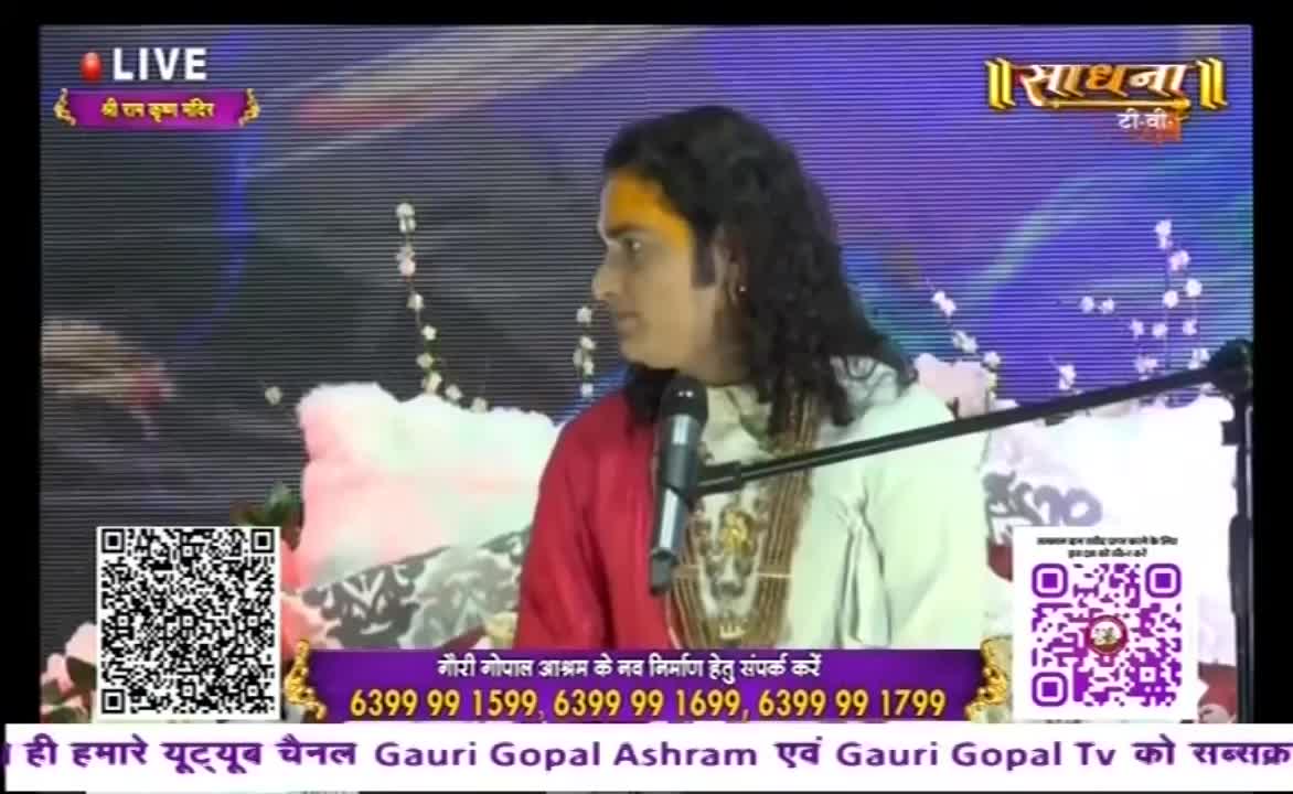 A man with long, dark hair and a red shawl speaks into a microphone, his gaze directed to the side. The Sadhna TV logo and a "LIVE" banner are visible, along with contact information for a temple.
