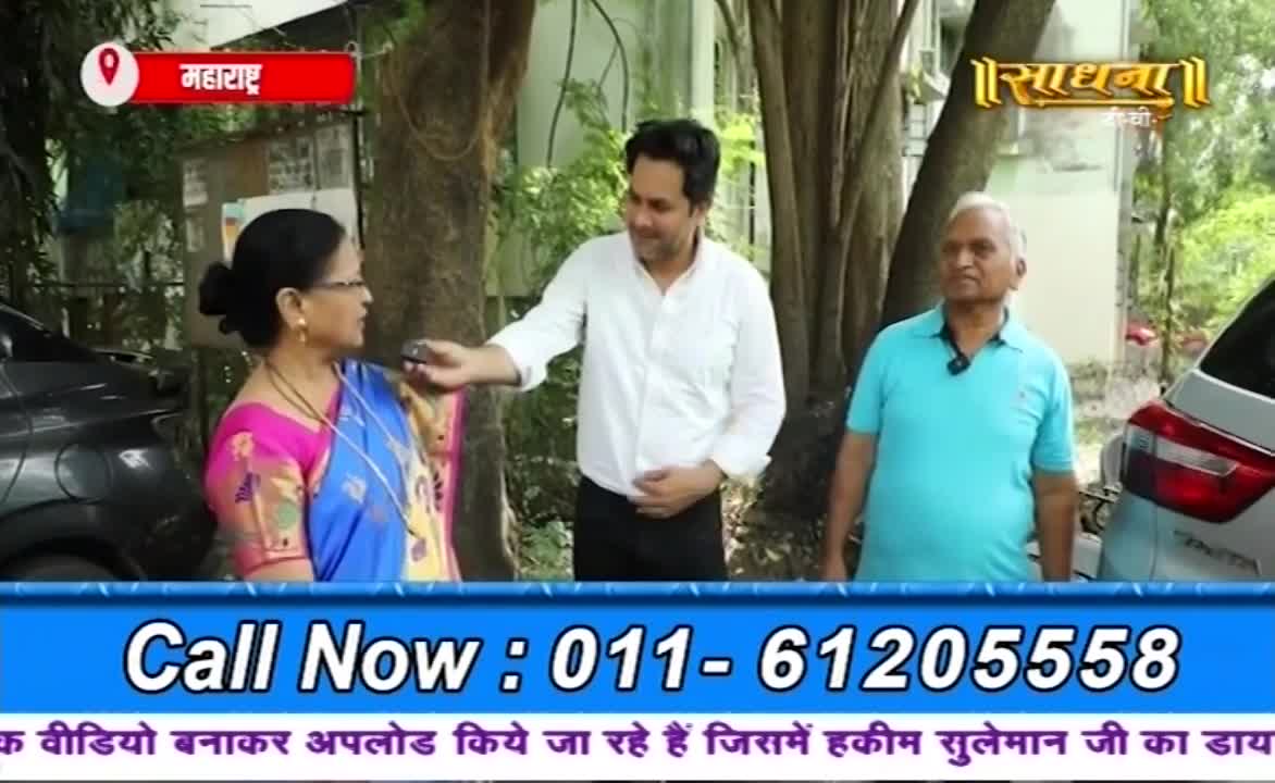 A man in a white shirt gestures toward a woman wearing a blue and pink sari, as if speaking to her. An older man in a turquoise shirt stands beside them, and the Sadhna logo is visible in the background.
