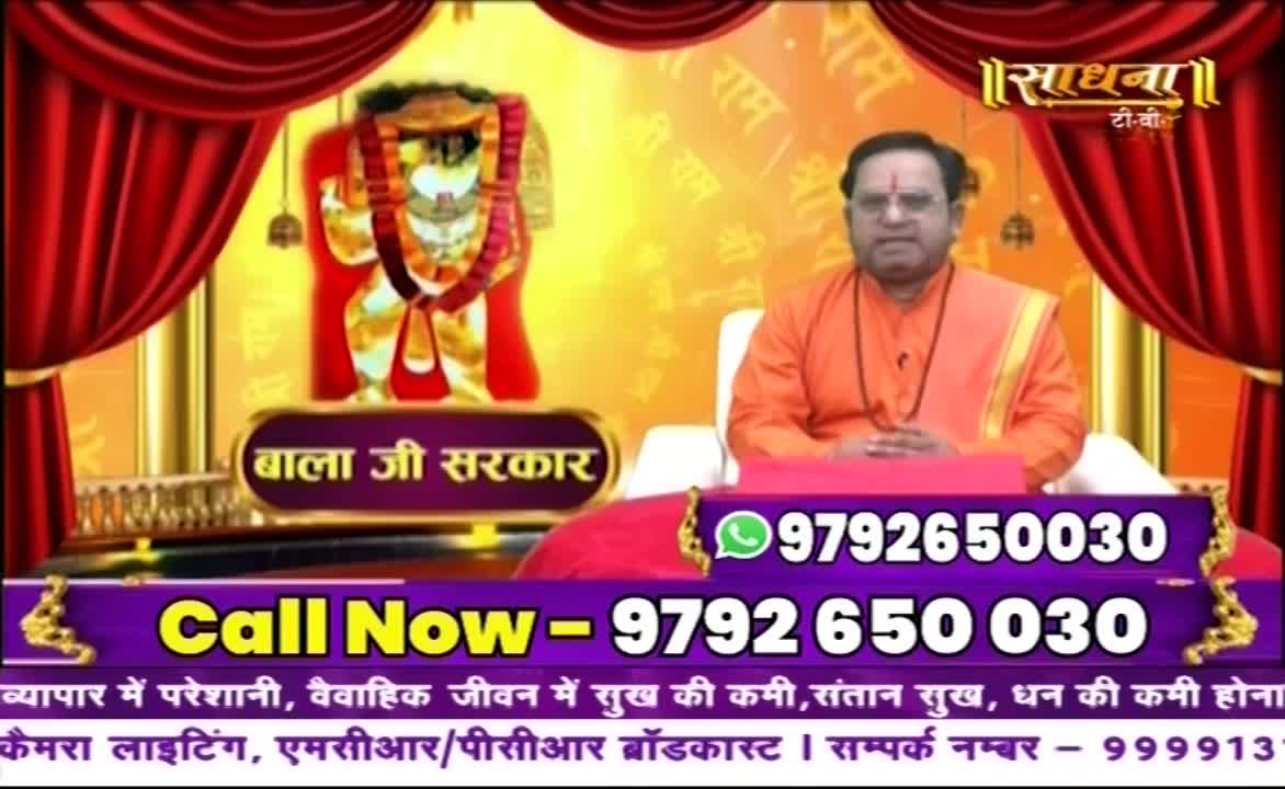 A man in orange robes sits before a backdrop featuring an image of a deity. A phone number flashes on the screen, inviting viewers to call.
