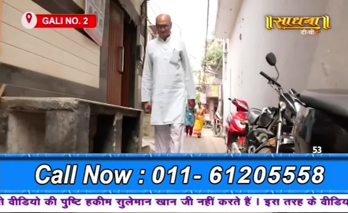 A man in white walks toward the camera down a narrow street, past parked motorbikes. Further down the alley, a woman in a yellow sari and others are walking away.
