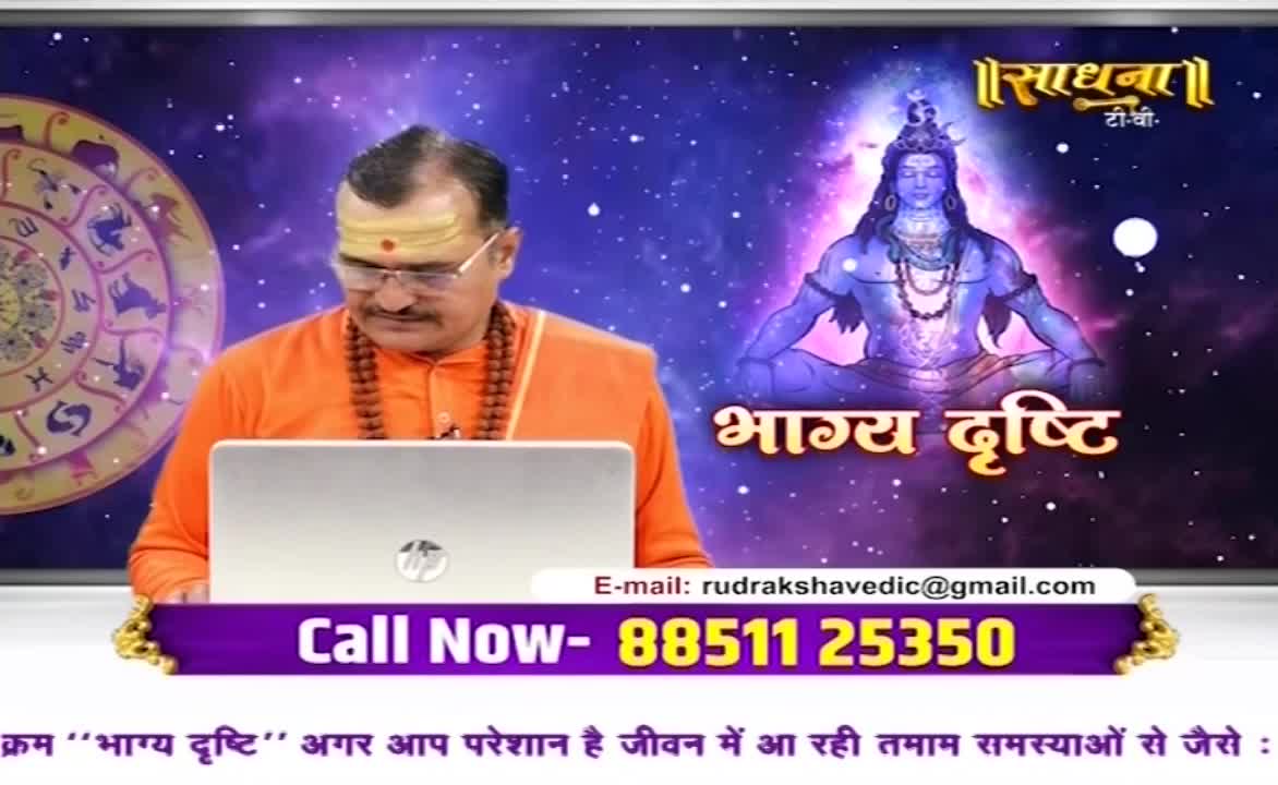 A man in saffron robes sits at a desk, looking down at a laptop. Behind him, a cosmic backdrop features an image of a deity and the Sadhna logo.
