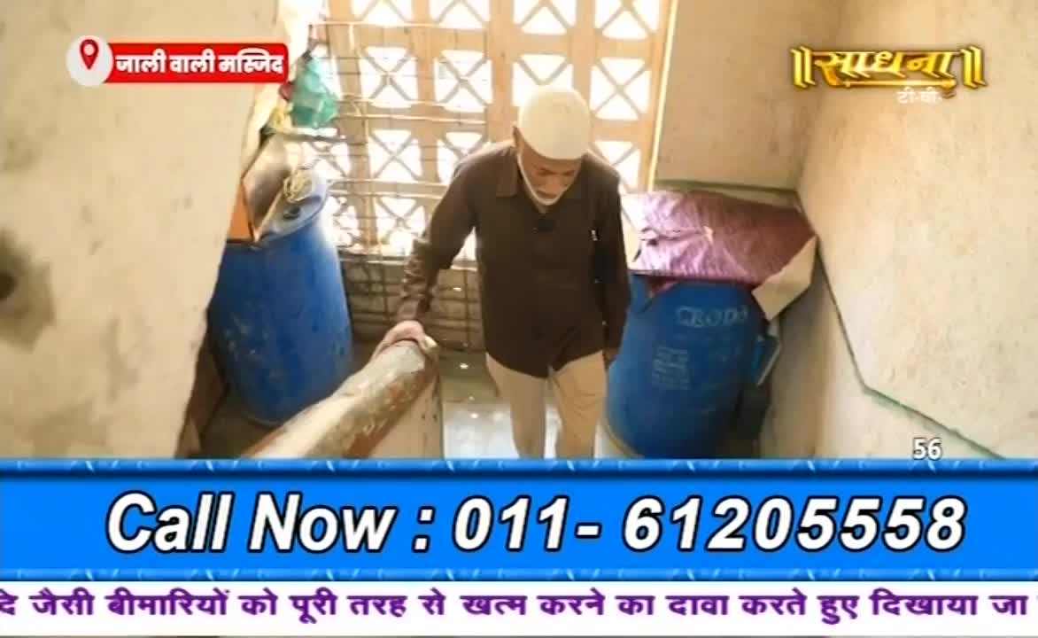 An older man in a white cap descends a narrow, cluttered staircase. He's holding onto a wooden railing, passing large blue barrels and what looks like Sadhna's camera crew, filming.
