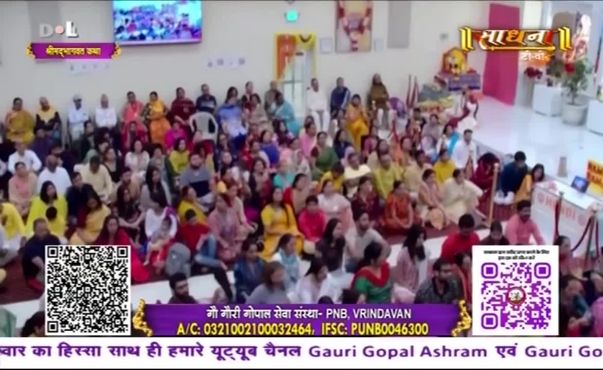 A large crowd sits on the floor, listening to something. The Sadhna TV logo is visible, and there's a QR code on the screen.
