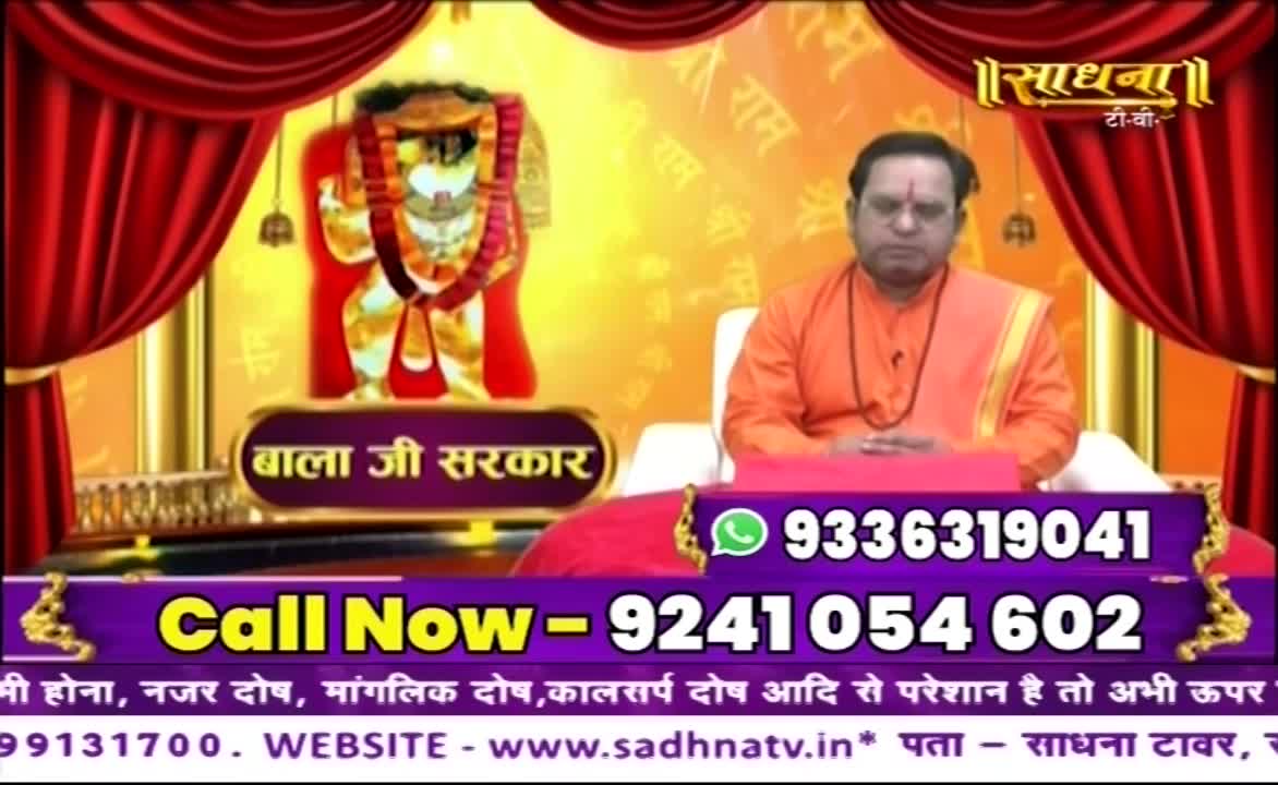 A man in orange robes sits on a red cushion, facing the camera. Behind him, a golden backdrop displays an image of a deity and the Sadhna TV logo, with phone numbers prominently displayed at the bottom.
