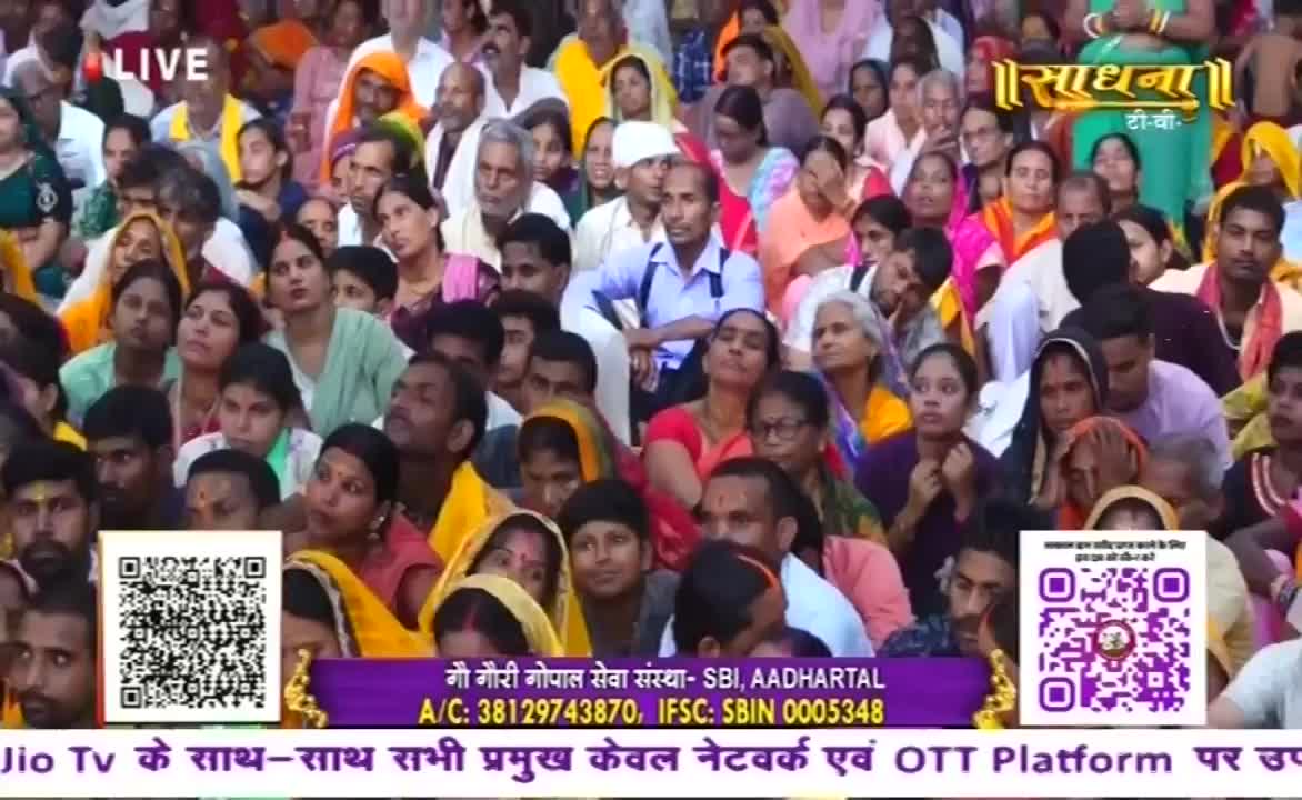 A large crowd, many in brightly colored saris and headscarves, looks upward, their attention focused on something just out of view. The broadcast from Sadhna TV is live, and the scene is likely from India.
