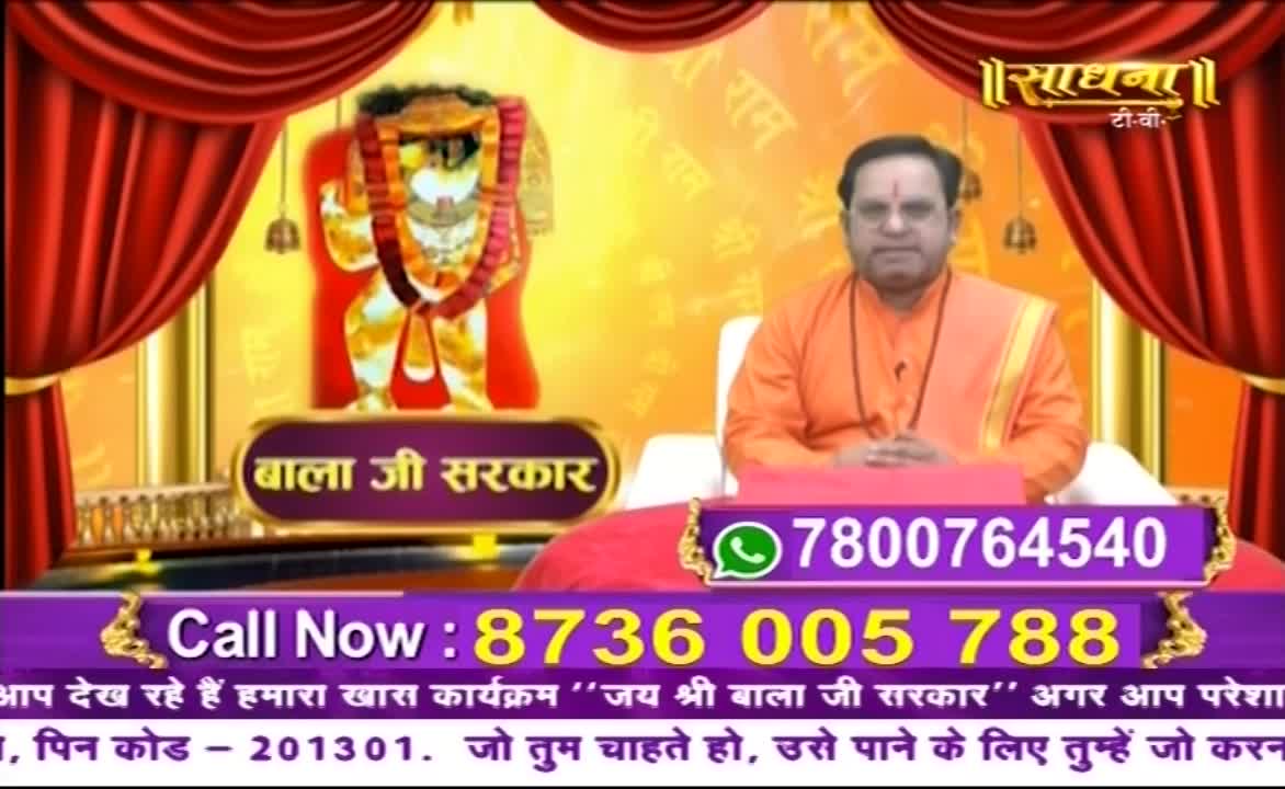 A man in saffron robes sits on a red cushion, facing the camera on what appears to be a Sadhna TV set. Behind him, a golden backdrop features an image of a deity, with phone numbers for viewers to call displayed prominently below.
