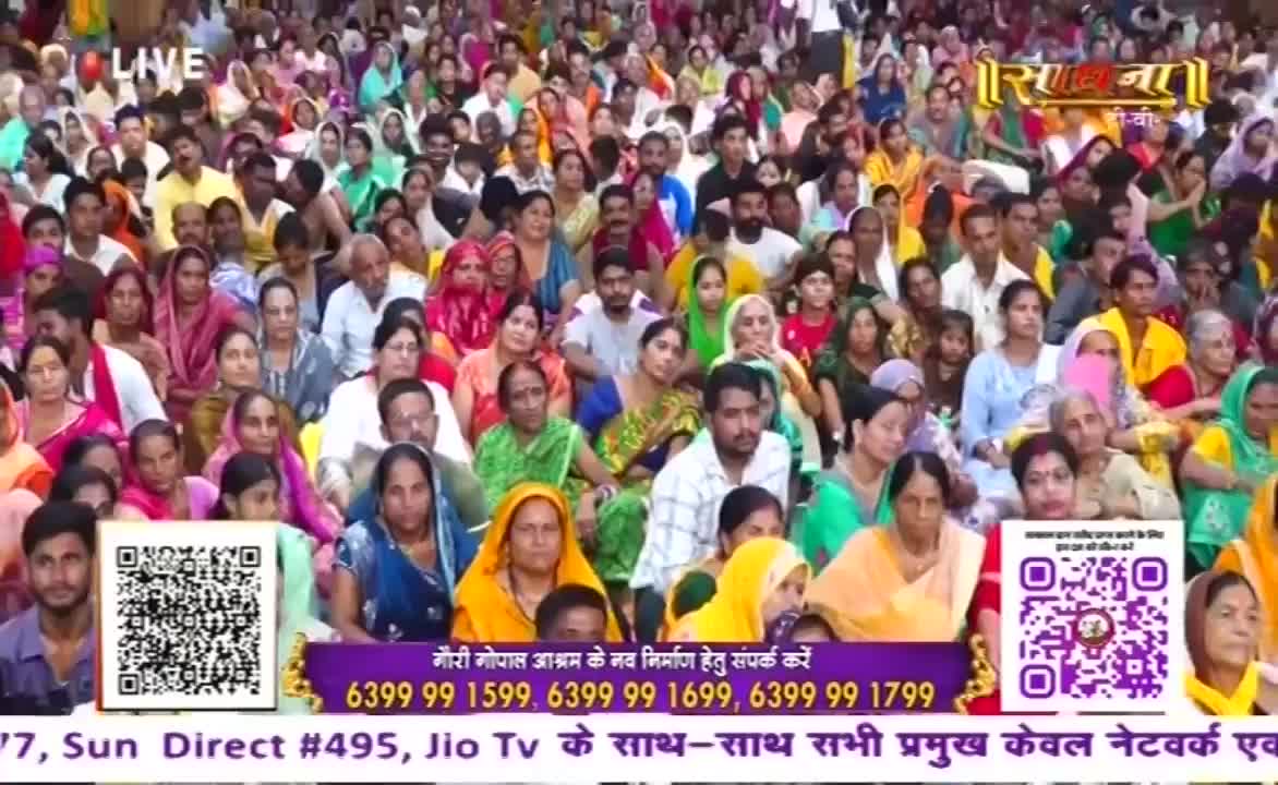 A large crowd, many in colorful saris and headscarves, sits closely together, facing the camera. The scene appears to be a live broadcast from India, possibly a Sadhna event, with text and QR codes in the foreground.

