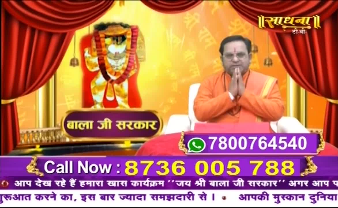 A man in orange robes sits with hands clasped, a backdrop of a deity behind him. The Sadhna TV logo and call-in numbers are displayed on the screen.
