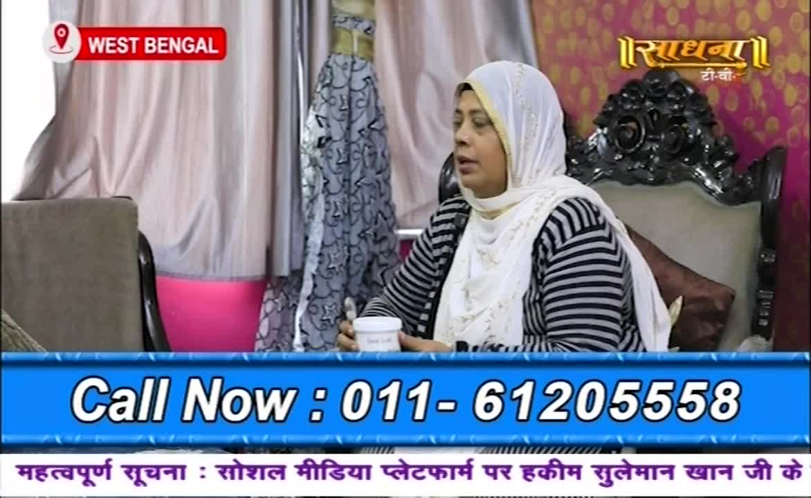 A woman in a white headscarf and striped sweater sits in a decorative chair, holding a small cup. The Sadhna TV logo is visible, and a phone number is displayed at the bottom of the screen.
