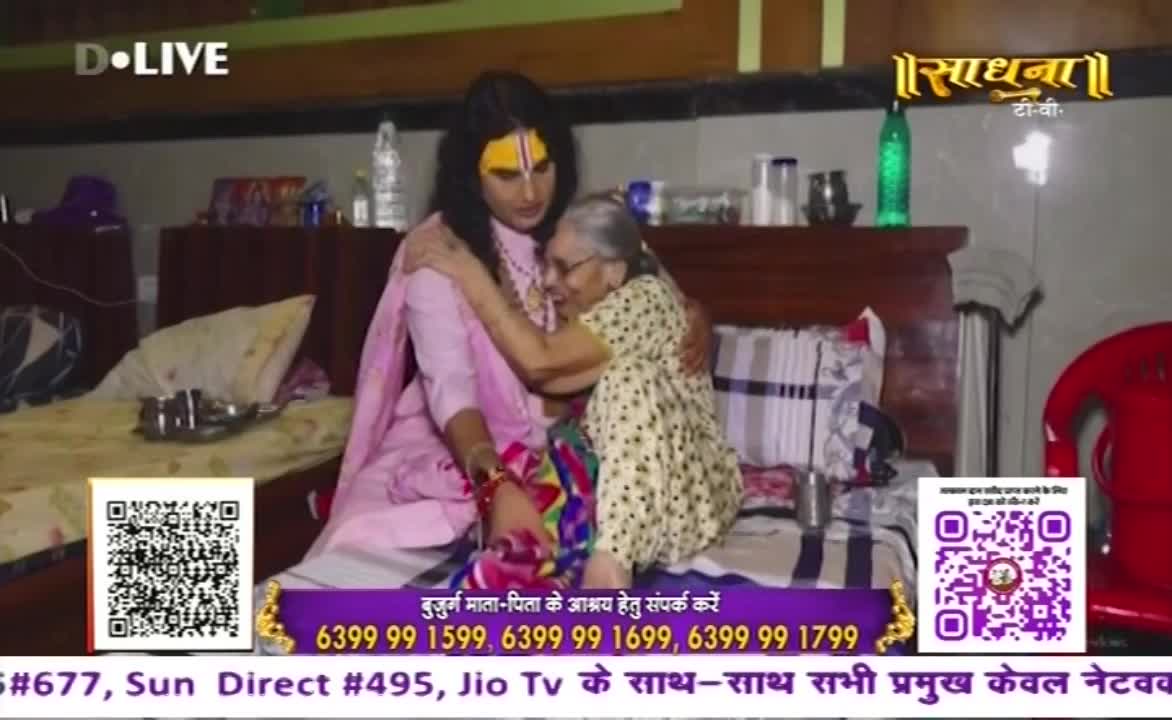 A person in a pink outfit and face paint embraces an elderly woman, both seated on a bed. The scene is framed by a television broadcast from "Sadhna" in India, with contact information displayed at the bottom.
