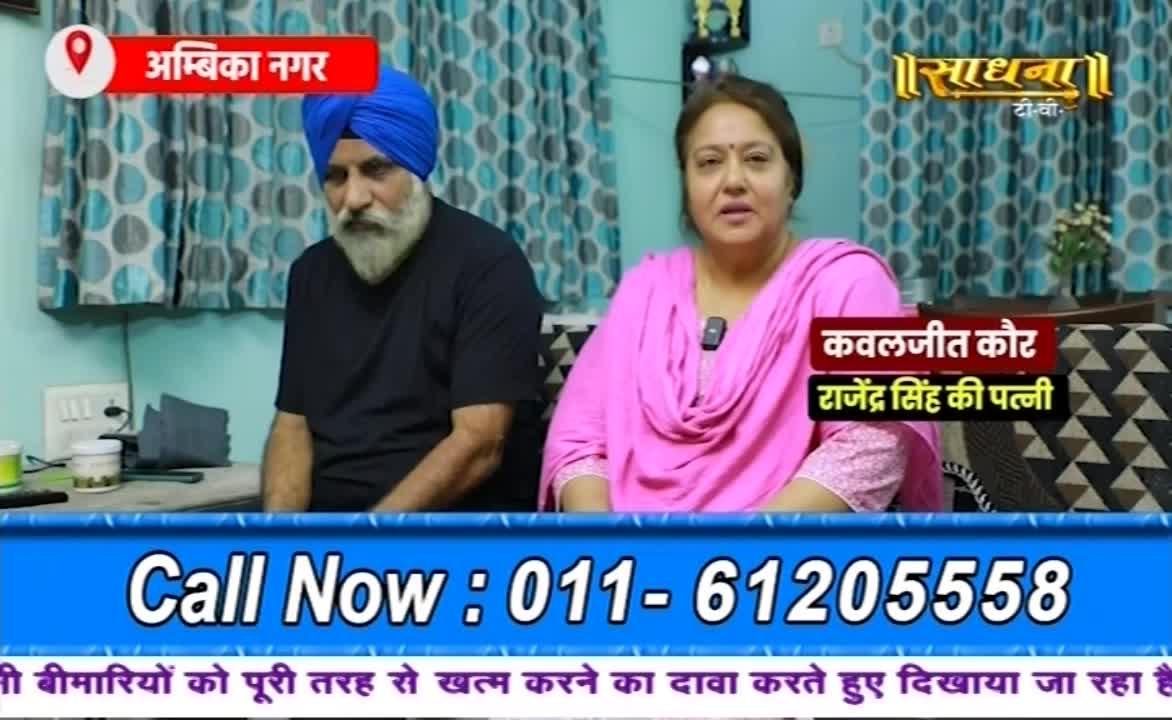 A man in a blue turban and a woman in a pink shawl sit side-by-side, speaking on a Sadhna TV program. A phone number flashes on the screen below them.
