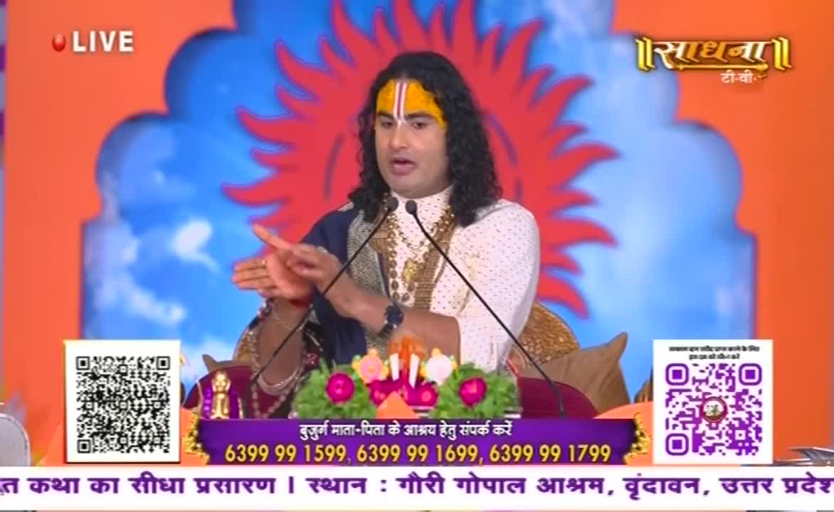 A man with long, dark hair and a painted forehead gestures with his hands while speaking into two microphones. The Sadhna TV logo and contact information for elder care are displayed below him.

