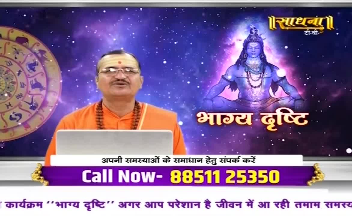 A man in saffron robes, likely in India, speaks earnestly towards a laptop, with a cosmic backdrop featuring an image of Shiva. The Sadhna TV logo and a phone number for immediate assistance are prominently displayed on the screen.
