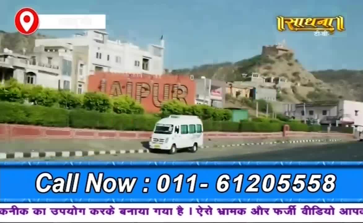 A white minibus drives past a large sign that says "JAIPUR" in bold letters. The Sadhna logo sits in the top right corner, and a phone number is displayed at the bottom.
