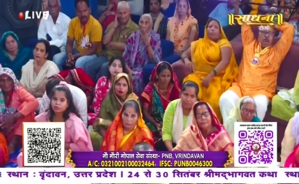 People sit close together, many women in colorful saris and headscarves, watching something off-camera. A man in an orange outfit raises his arms, and the Sadhna logo is displayed on the screen.
