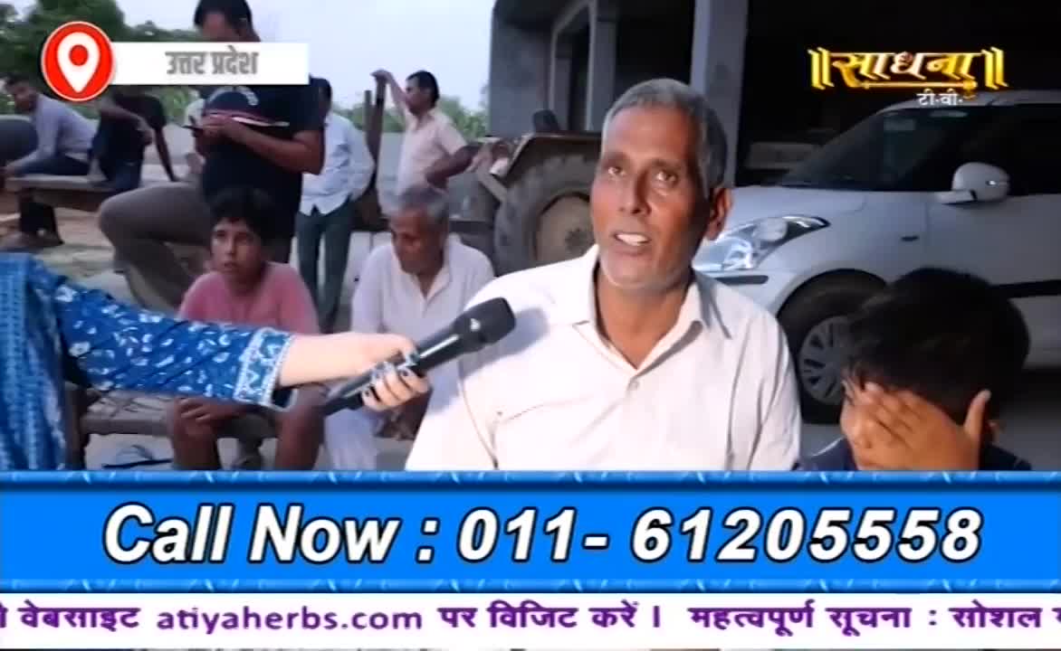 A man in a white shirt speaks into a microphone held by someone out of frame, while the Sadhna TV logo is visible. Behind him, a group of people observe, and a boy covers his face with his hands.
