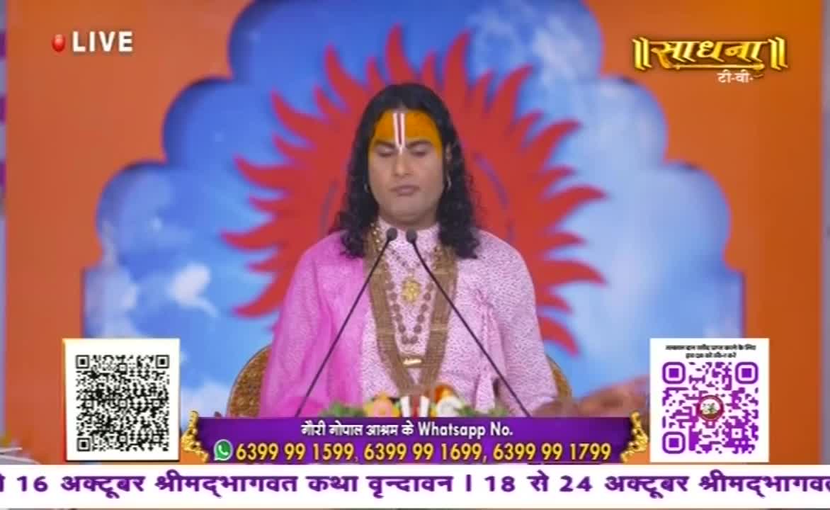 A man with a prominent forehead marking stands at a podium, speaking into two microphones. Behind him, the Sadhna TV logo and a stylized sunburst decorate the backdrop.
