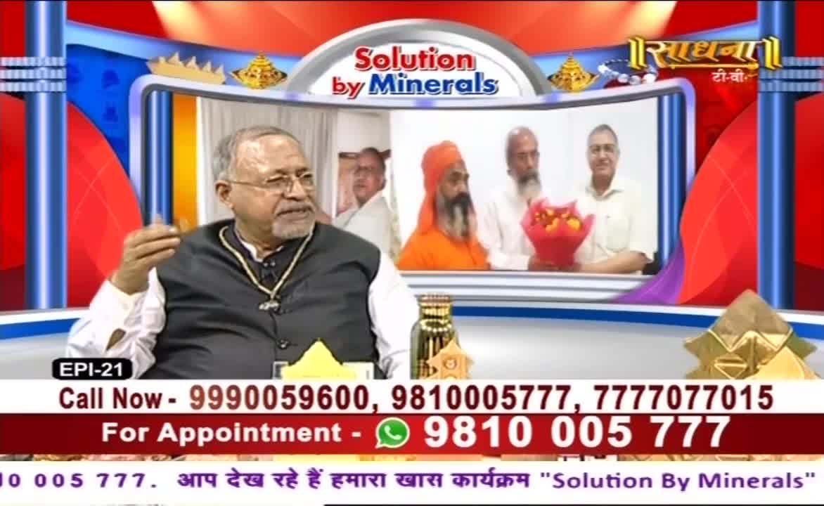 A man in a dark vest gestures with his hand as he speaks on a Sadhna TV show. Behind him, a screen displays other men, one holding a bouquet of flowers.
