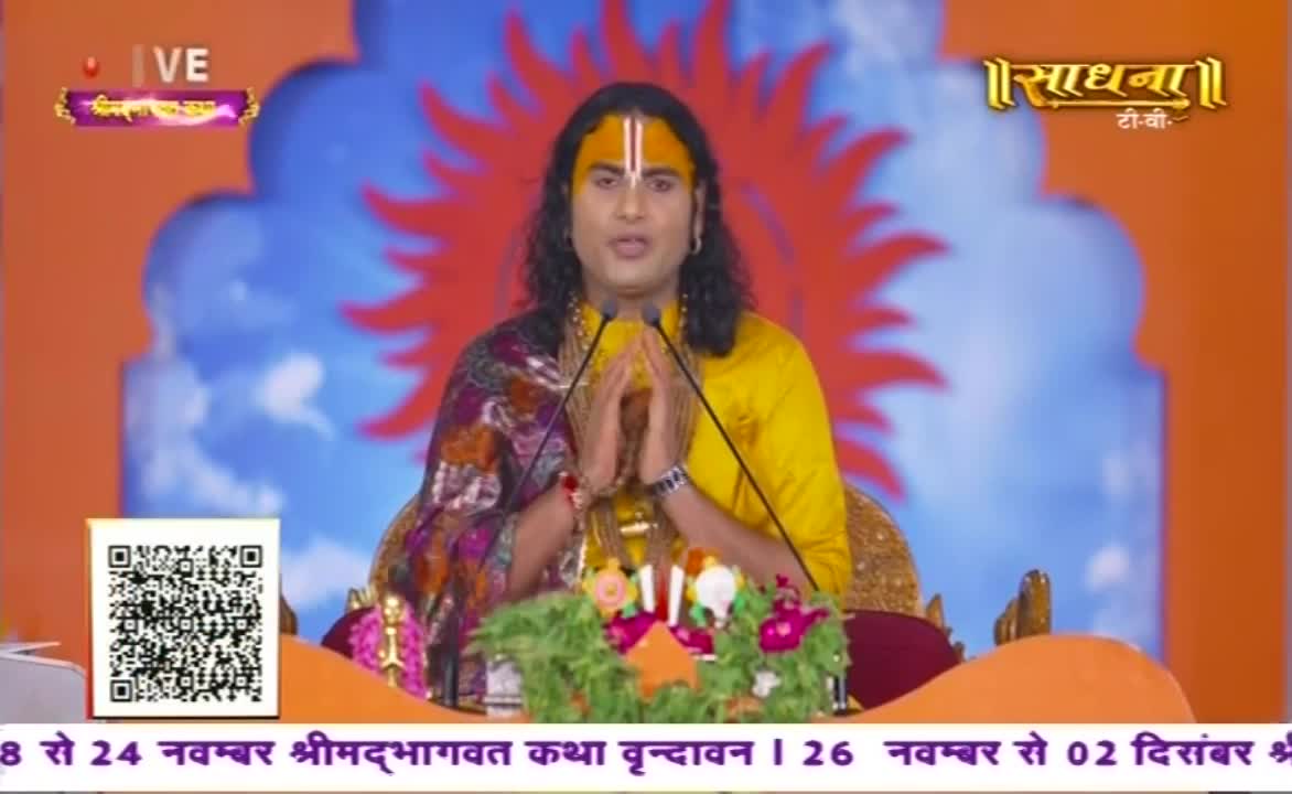 A man in a yellow shirt, with a painted forehead, speaks into two microphones, hands clasped in front of him. The Sadhna TV logo is visible, and the background is a stylized, colorful design.
