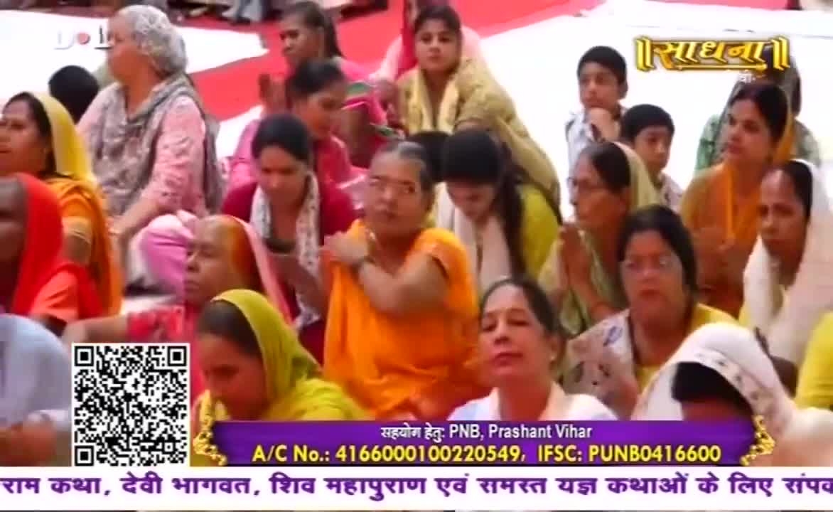 A crowd of women, many wearing headscarves, are seated on the floor, some with their hands clasped. The Sadhna logo is visible, and a banner displays information about donations.
