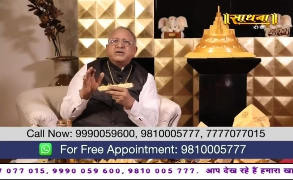 A man in a black vest gestures with his hands while holding a small, golden object. The Sadhna TV logo sits above him, and a phone number scrolls across the bottom of the screen.
