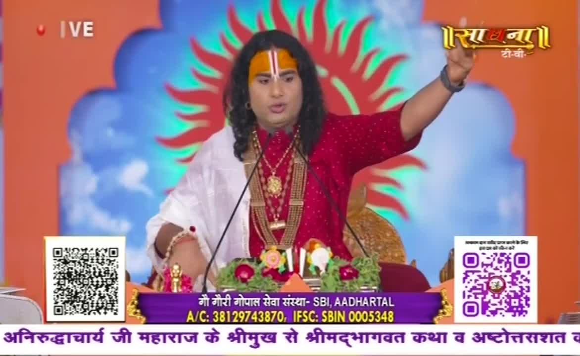 A man in a red shirt with a white shawl gestures with his right hand, speaking into a microphone. The Sadhna TV logo is visible in the top right corner, and below, a banner advertises a live broadcast from India.
