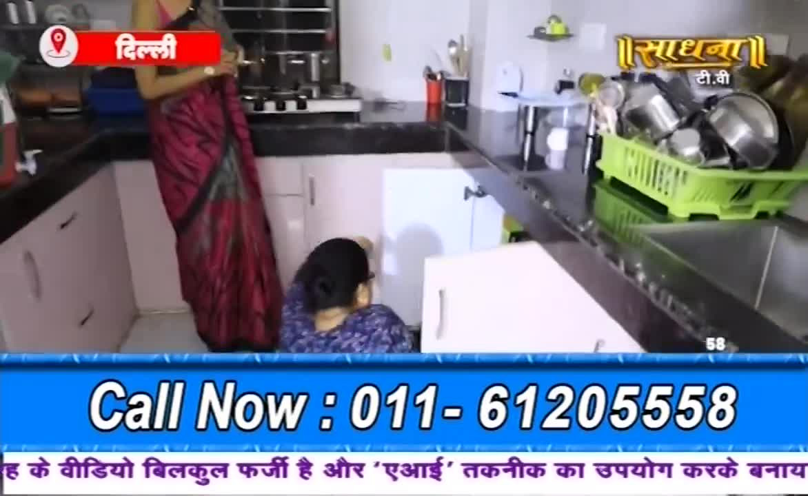 In a kitchen setting, a woman in a maroon and black sari stands near a cabinet while another woman crouches, seemingly focused on something inside. The Sadhna TV logo and a phone number are displayed at the bottom of the screen.
