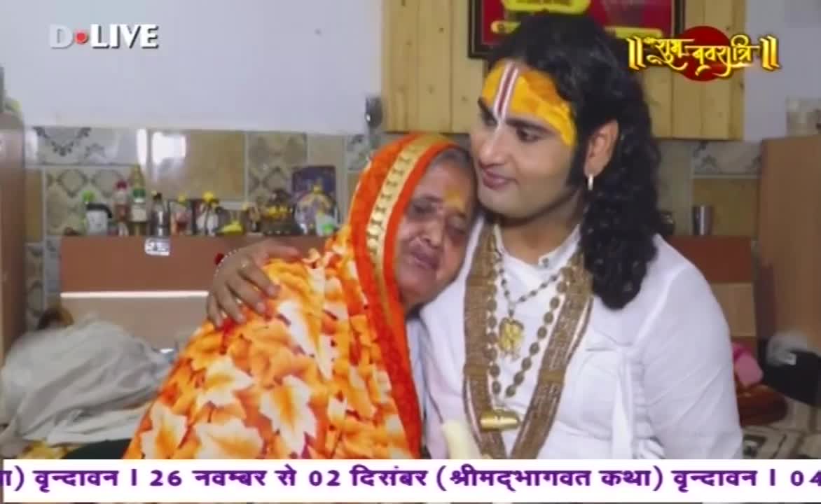 A man with a painted forehead and long hair embraces an older woman, both close together. She wears a bright orange and yellow headscarf, and he has a necklace with a large pendant.
