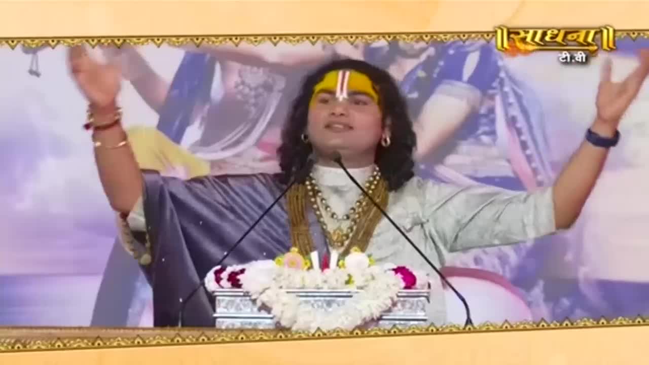 A man with a yellow mark on his forehead speaks into two microphones, his arms raised. He's wearing a light-colored shirt, and a garland rests on the table in front of him.
