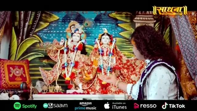 A man with long, dark hair gestures toward two ornate statues, adorned with garlands and elaborate clothing. The Sadhna logo sits in the top right corner, and the scene is set with brightly painted backdrops.
