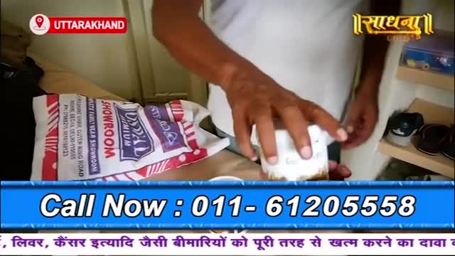 A man holds a small white container, possibly a product being advertised on the Sadhna channel. A shopping bag from a local showroom rests nearby, and a phone number is displayed on the screen.
